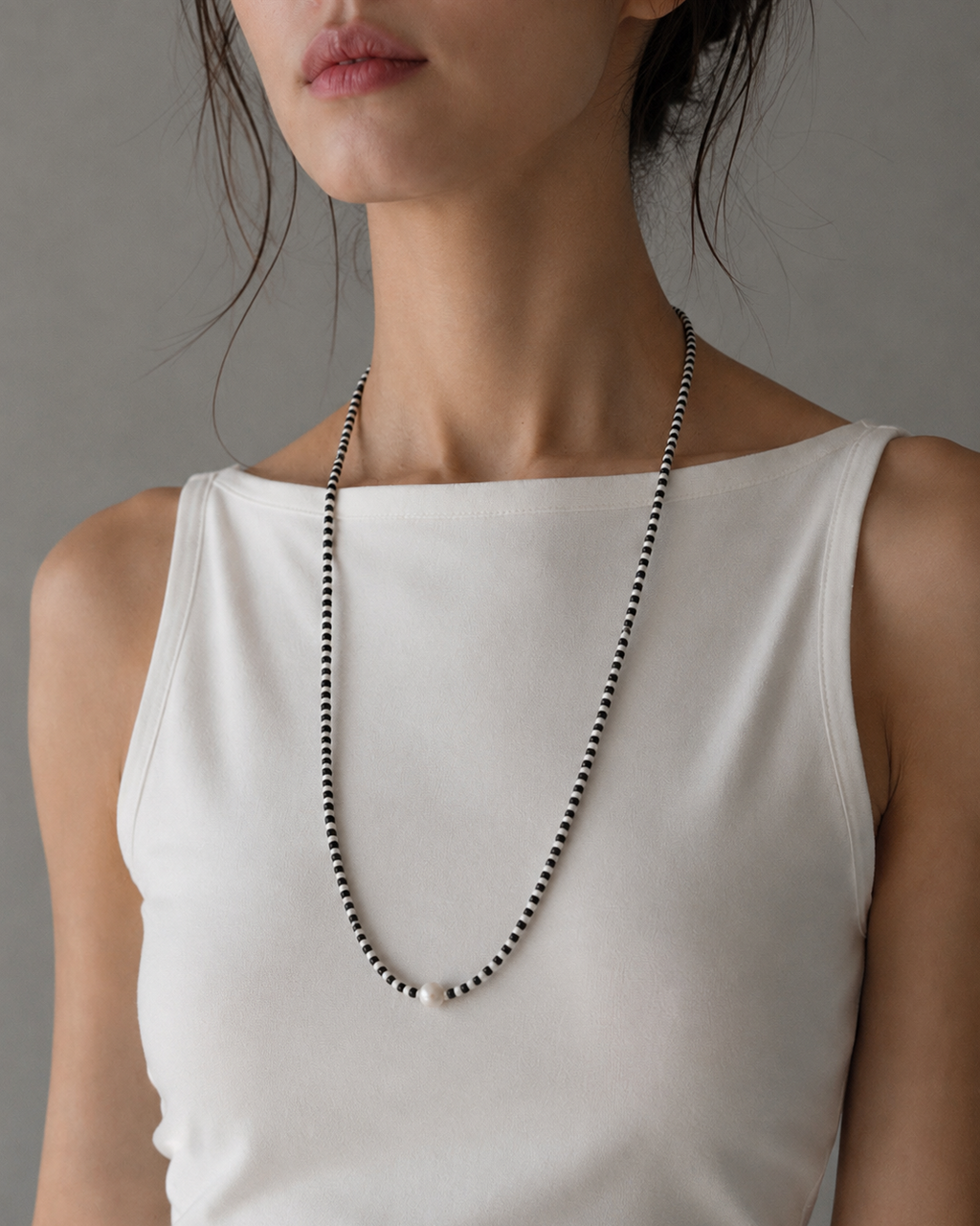 A close-up of a model wearing a long, delicate beaded necklace with alternating black and white beads, styled with a white sleeveless top against a neutral gray background.