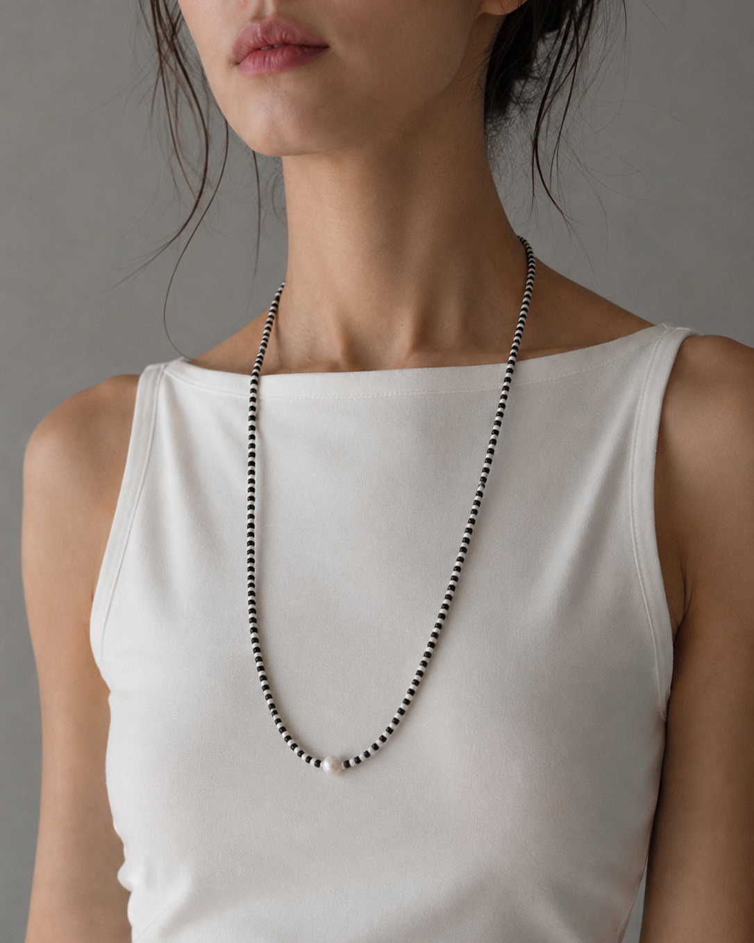 A close-up of a model wearing a long, delicate beaded necklace with alternating black and white beads, styled with a white sleeveless top against a neutral gray background.