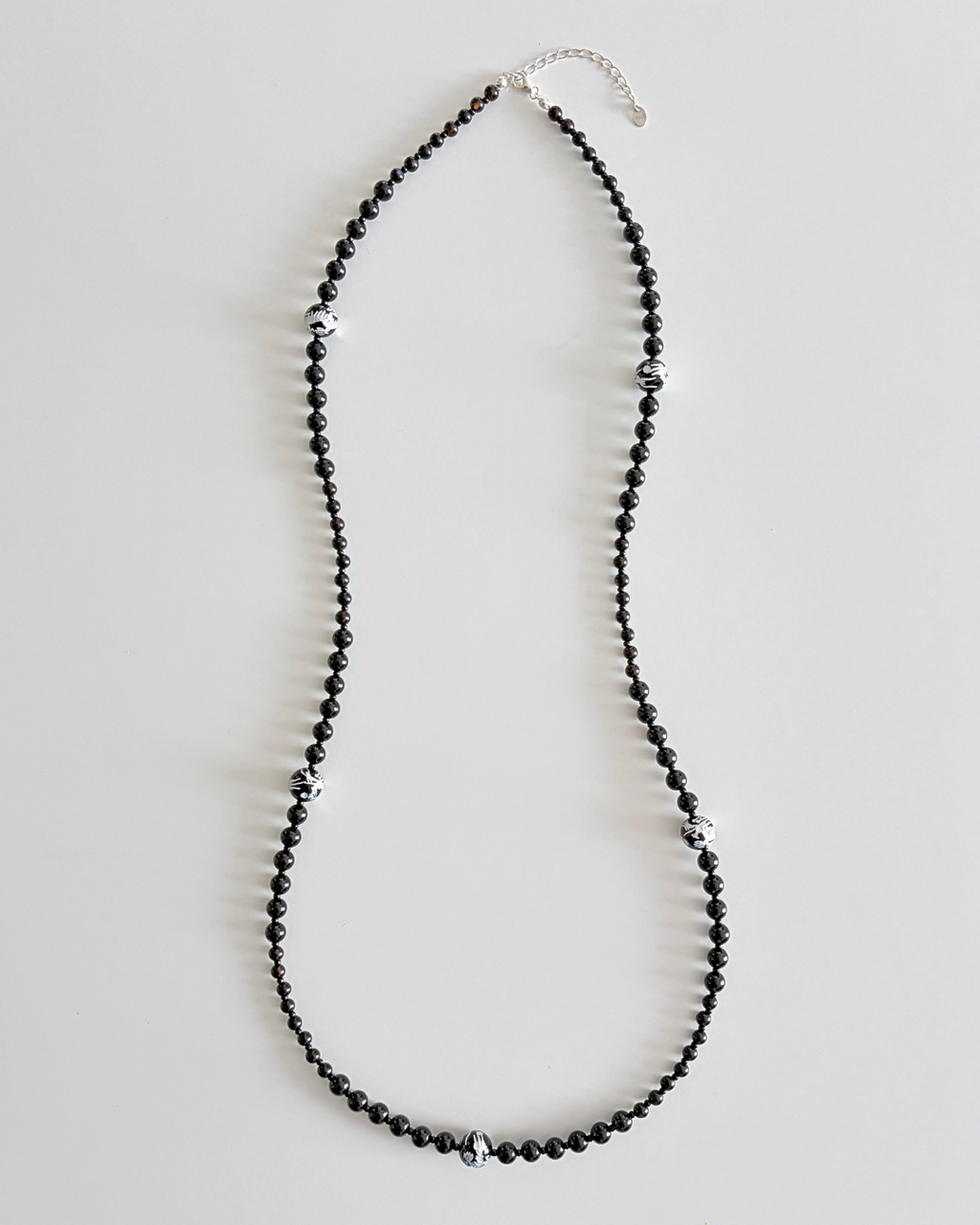 A long beaded necklace made of small black faceted beads with scattered silver-toned spacer beads and a silver clasp, displayed on a light gray background.