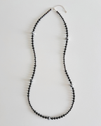 A long beaded necklace made of small black faceted beads with scattered silver-toned spacer beads and a silver clasp, displayed on a light gray background.