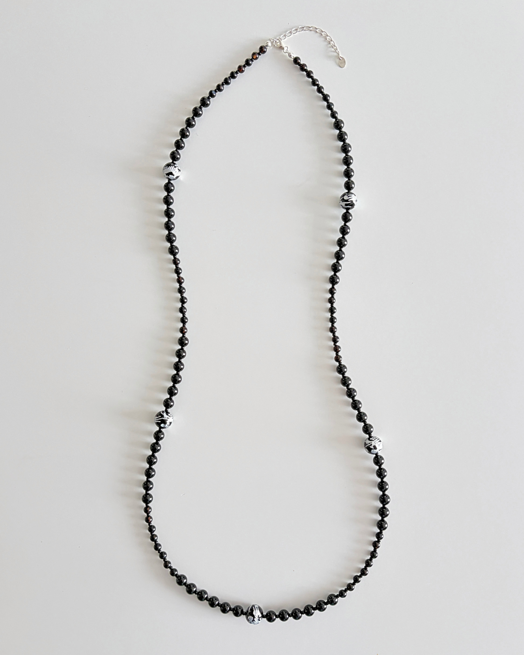 A long beaded necklace made of small black faceted beads with scattered silver-toned spacer beads and a silver clasp, displayed on a light gray background.