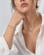 A close-up lifestyle shot of a model wearing a silver chain necklace with a small square pendant and a matching silver bracelet, styled with a white button-down shirt against a light gray background.