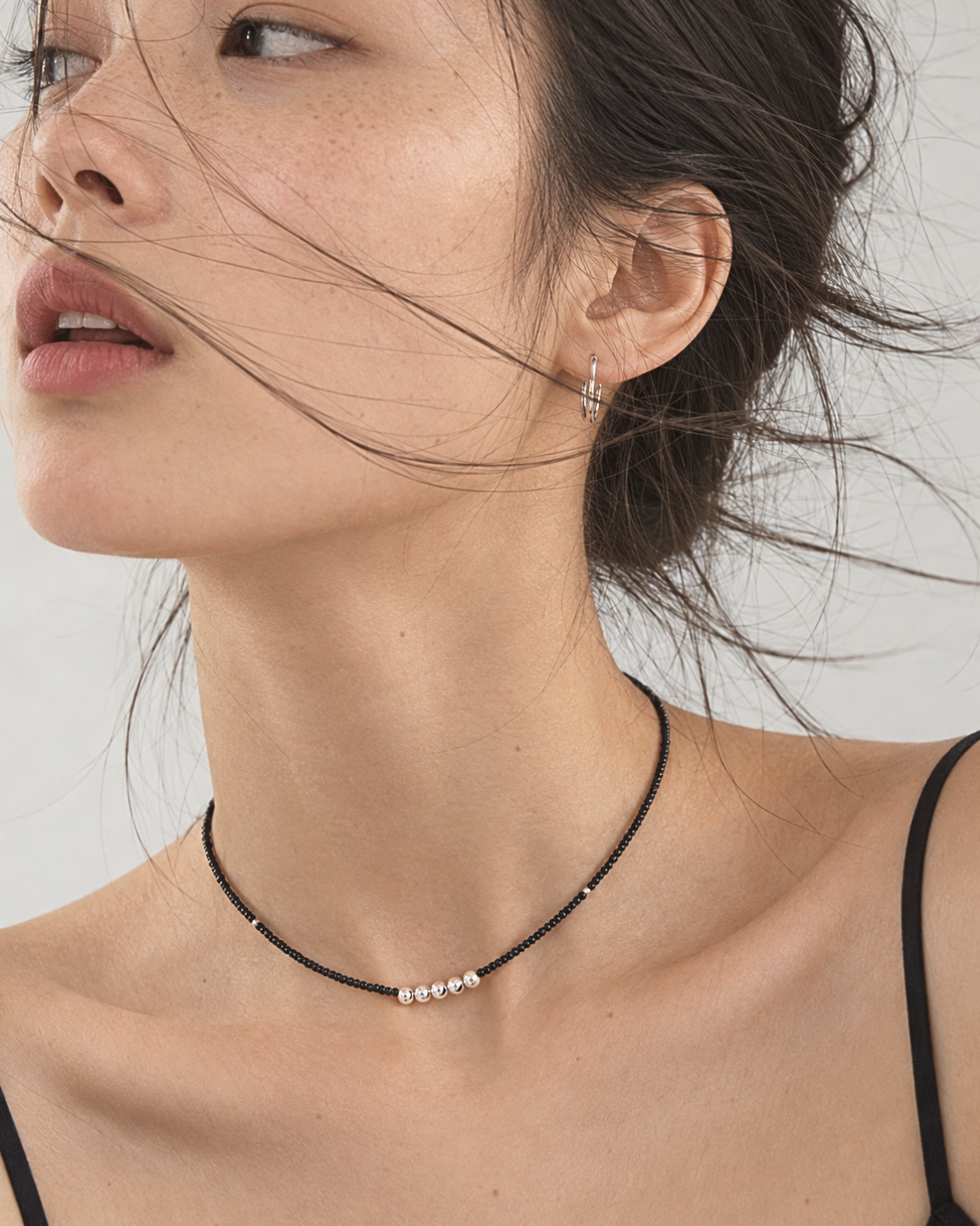 A close-up lifestyle shot of a model wearing a delicate black beaded choker necklace with a small silver bead cluster at the front, paired with a silver hoop earring.