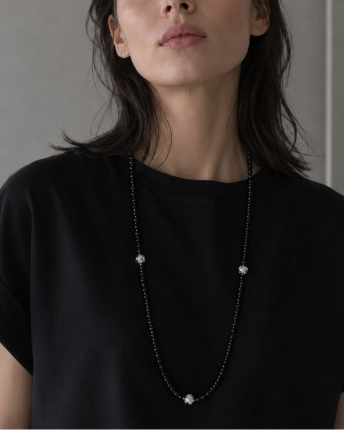 A woman with dark hair wearing a black t-shirt and a long silver beaded necklace with small white bead accents, photographed against a neutral gray wall.
