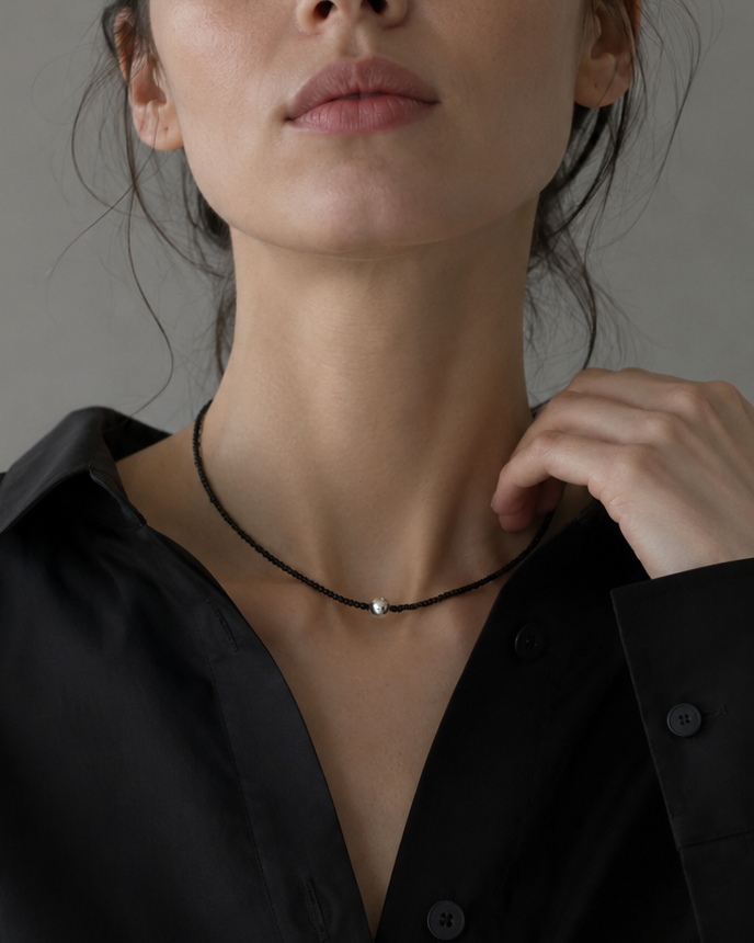 A close-up of a model wearing a delicate black cord necklace with a small silver bead pendant, paired with a black button-down shirt against a neutral gray background.