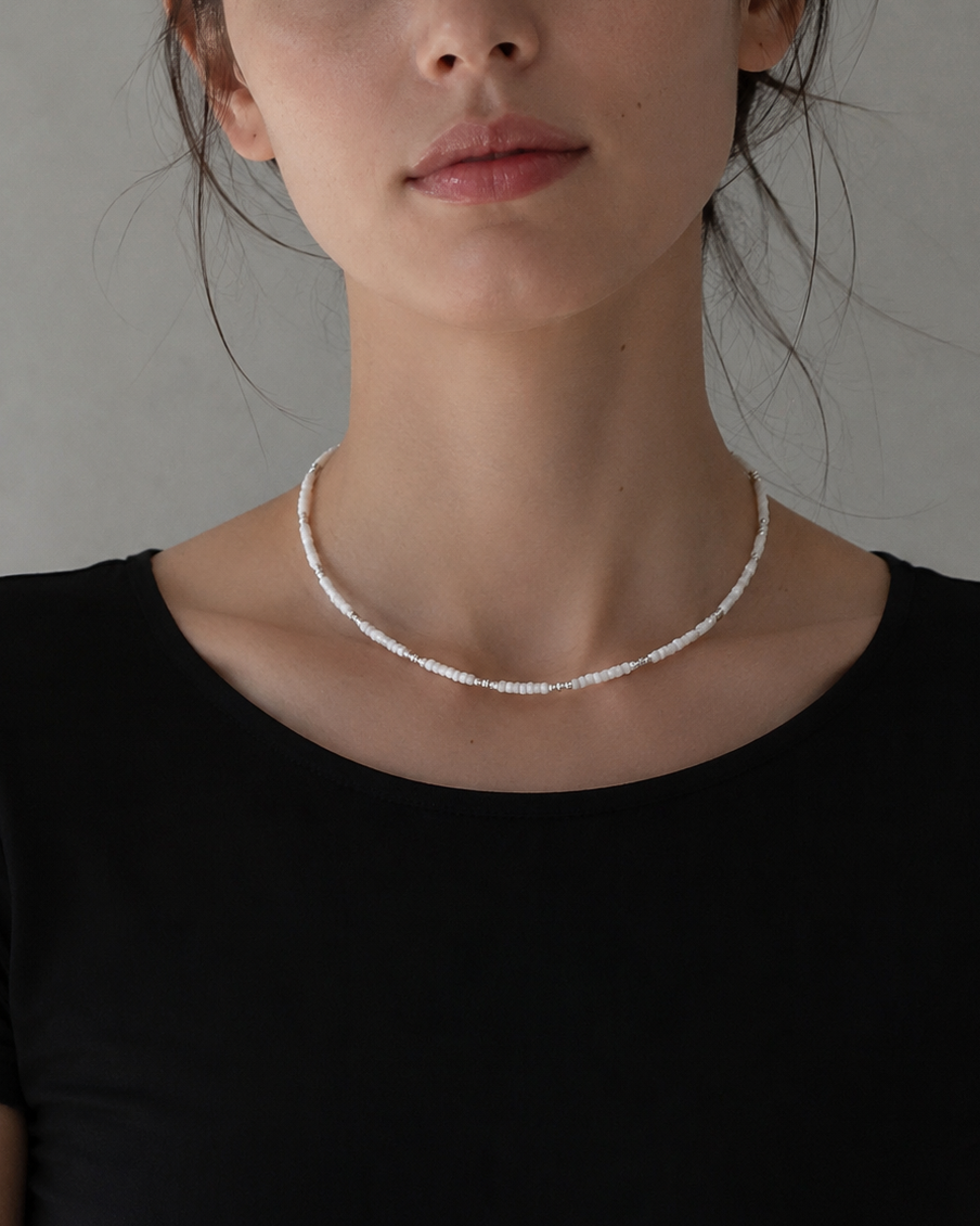 A woman wearing a delicate silver beaded necklace with a black top, photographed against a neutral gray background.