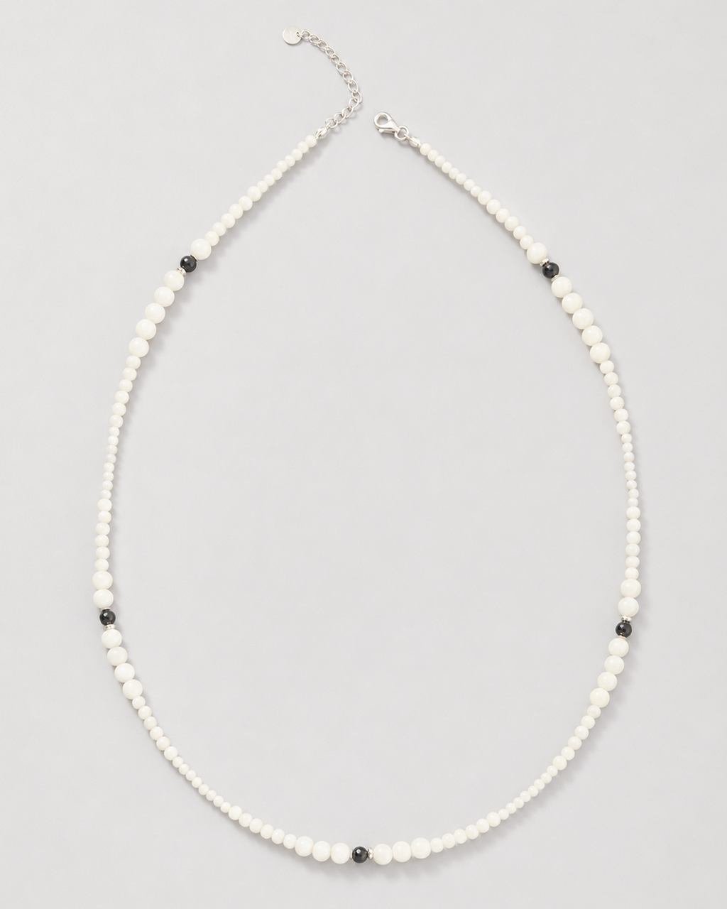 A delicate beaded necklace featuring small white beads with alternating tiny black beads and a silver-tone clasp, displayed on a neutral light gray background.