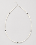 A delicate beaded necklace featuring small white beads with alternating tiny black beads and a silver-tone clasp, displayed on a neutral light gray background.