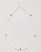 A delicate beaded necklace featuring small white beads with alternating tiny black beads and a silver-tone clasp, displayed on a neutral light gray background.