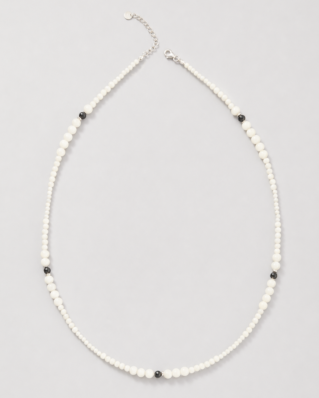 A delicate beaded necklace featuring small white beads with alternating tiny black beads and a silver-tone clasp, displayed on a neutral light gray background.