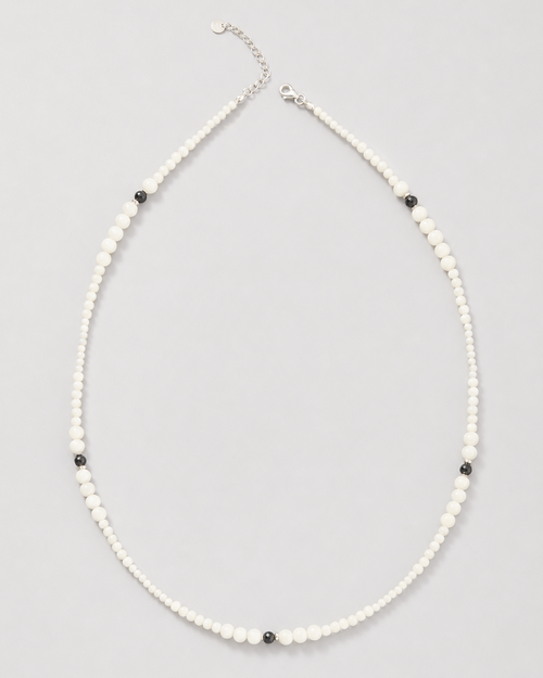 A delicate beaded necklace featuring small white beads with alternating tiny black beads and a silver-tone clasp, displayed on a neutral light gray background.