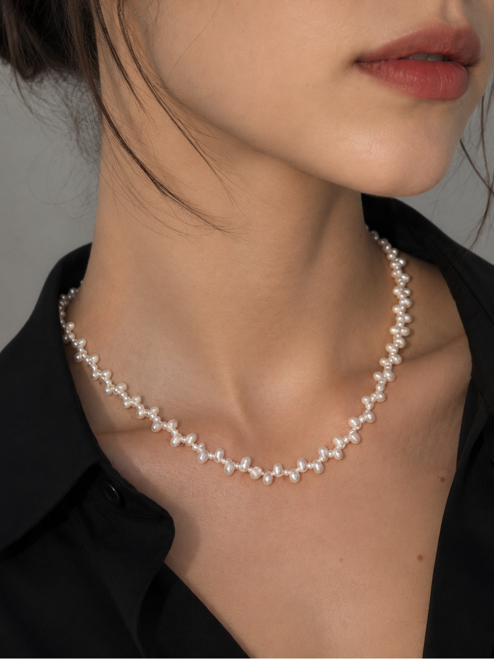 A close-up of a woman wearing a delicate pearl necklace with small white floral clusters on a silver-tone chain, paired with a black collared shirt against a neutral gray background.