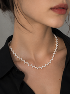 A close-up of a woman wearing a delicate pearl necklace with small white floral clusters on a silver-tone chain, paired with a black collared shirt against a neutral gray background.