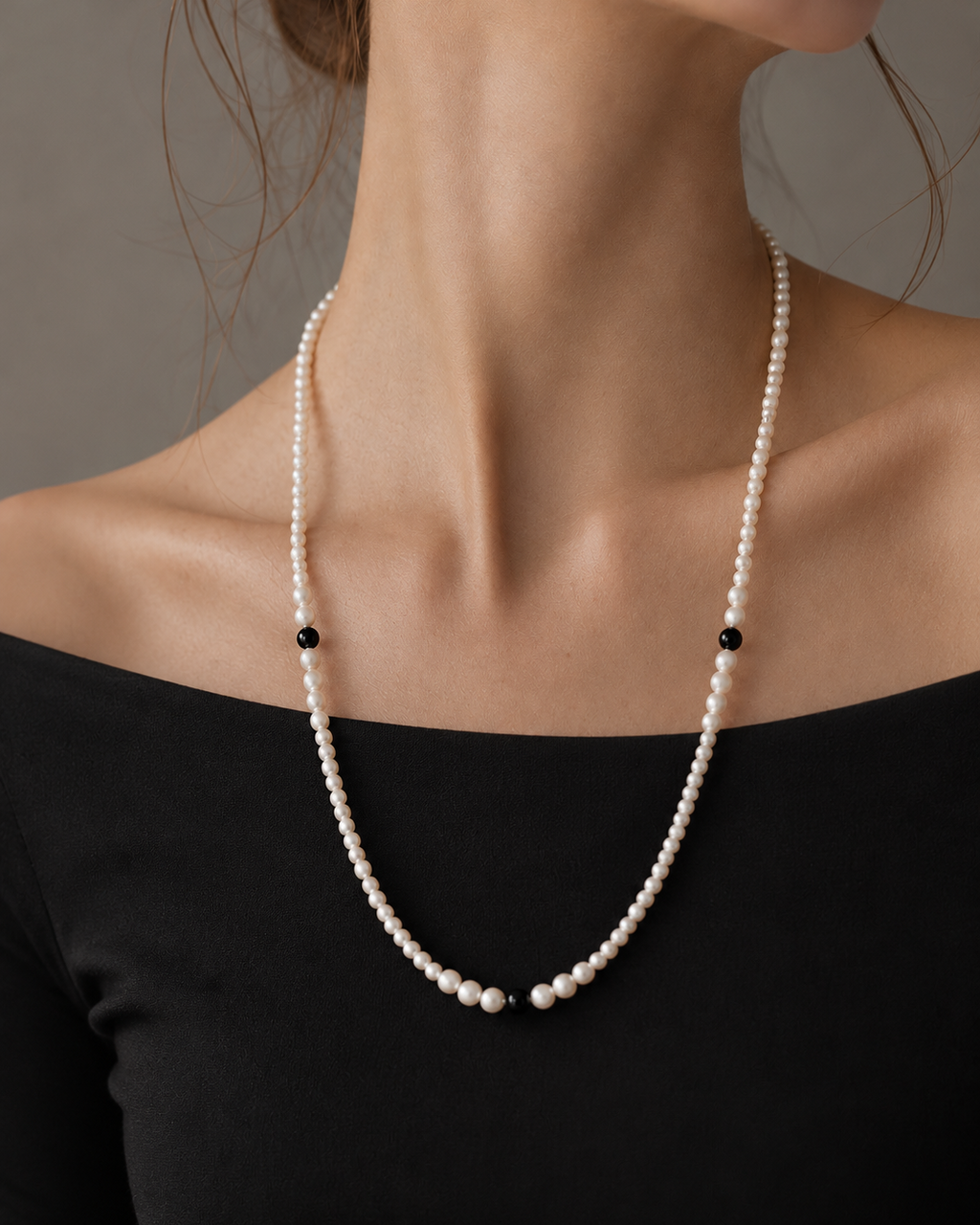 A close-up of a model wearing a delicate pearl necklace with small black bead accents, paired with a black off-the-shoulder top against a neutral gray background.