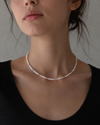 A close-up of a model wearing a delicate silver beaded choker necklace, paired with a black scoop-neck top against a neutral gray background.