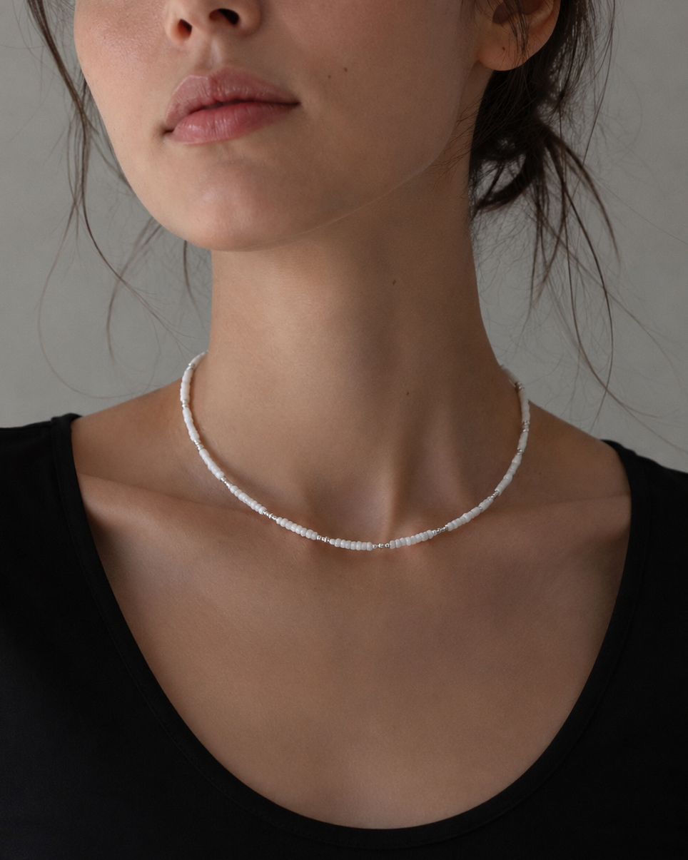 A close-up of a model wearing a delicate silver beaded choker necklace, paired with a black scoop-neck top against a neutral gray background.