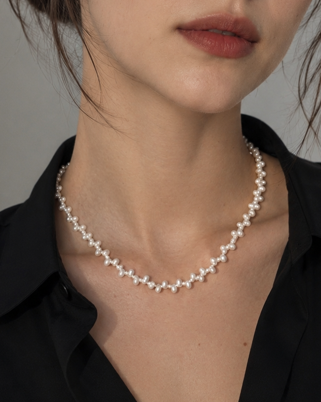 A close-up portrait of a woman wearing a delicate silver-toned necklace with small star-shaped charms, paired with a black collared shirt against a soft gray background.