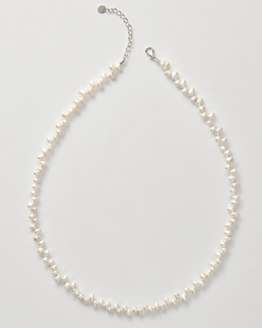 A delicate necklace made of small irregular white pearls on a silver-tone chain with an extender clasp, photographed on a light gray background.