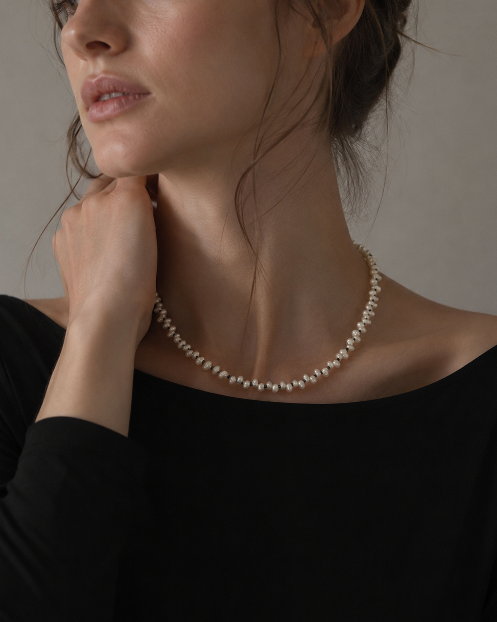 A close-up of a woman wearing a delicate pearl necklace with a silver-toned chain, paired with a black top against a neutral background.