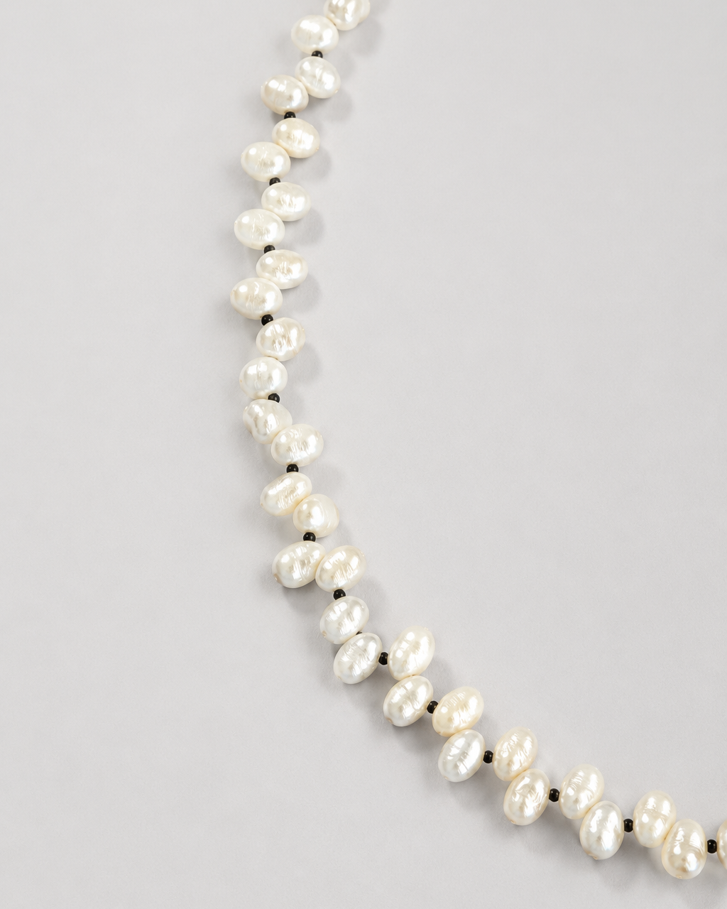 A strand of irregular white baroque pearls separated by small dark spacer beads, laid out on a light gray background.